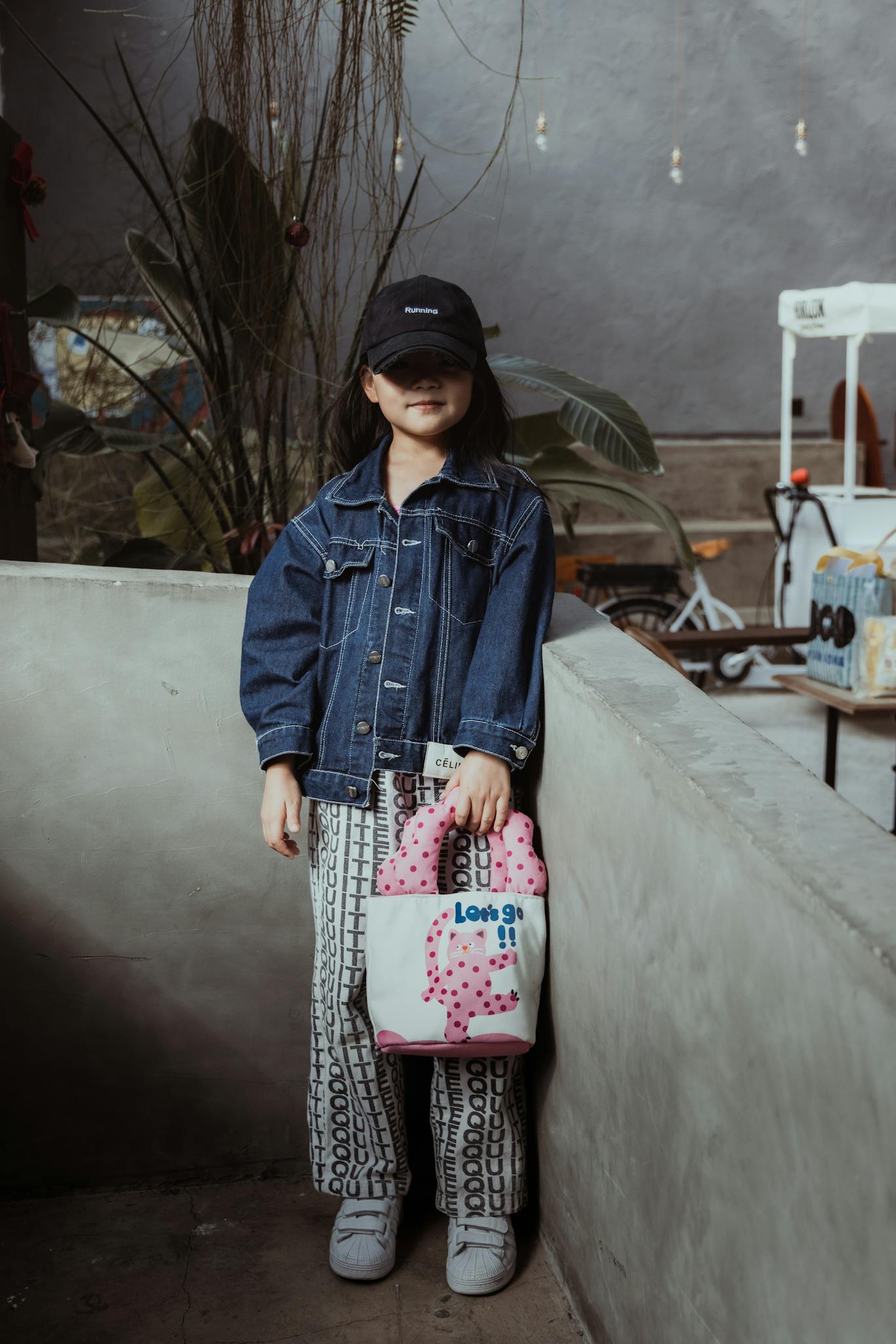 A fashionable child in a denim jacket and cap holding a cute tote bag outdoors.